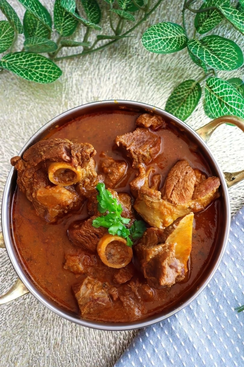 Close-up of a metal bowl filled with rich, reddish-brown lamb curry garnished with fresh cilantro.