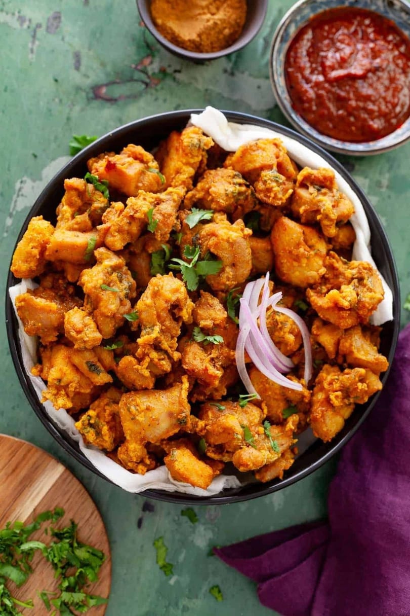 A black bowl filled with golden-brown fried chicken pakora, garnished with red onion and cilantro.