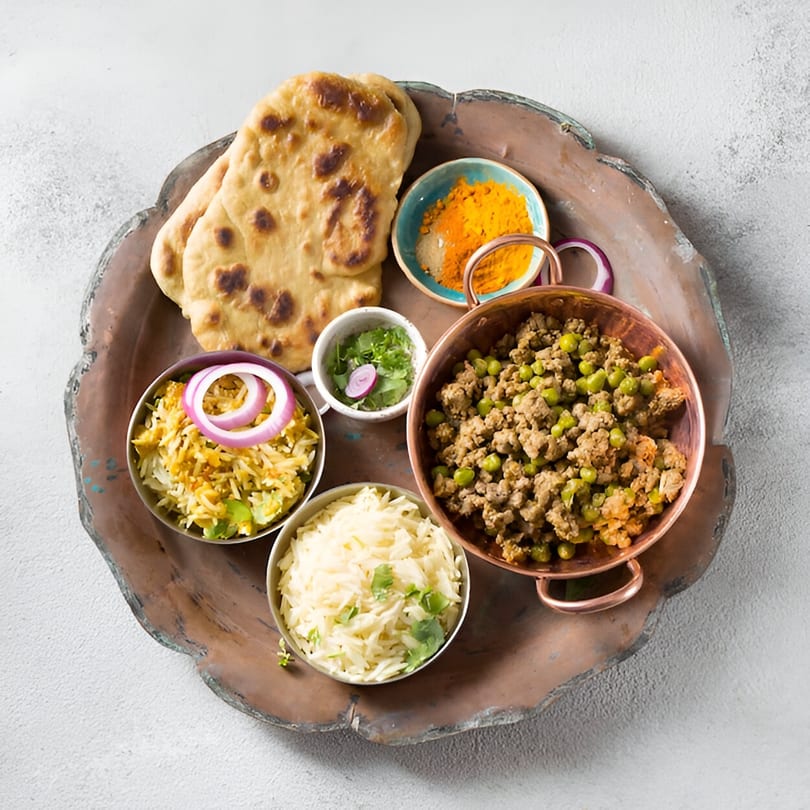 Indian meal with naan, keema matar, two rice dishes, and spices on a copper tray.