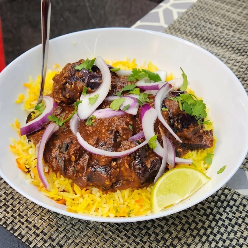 A white bowl filled with yellow rice, a rich brown stew, red onions, cilantro, and a lime wedge.