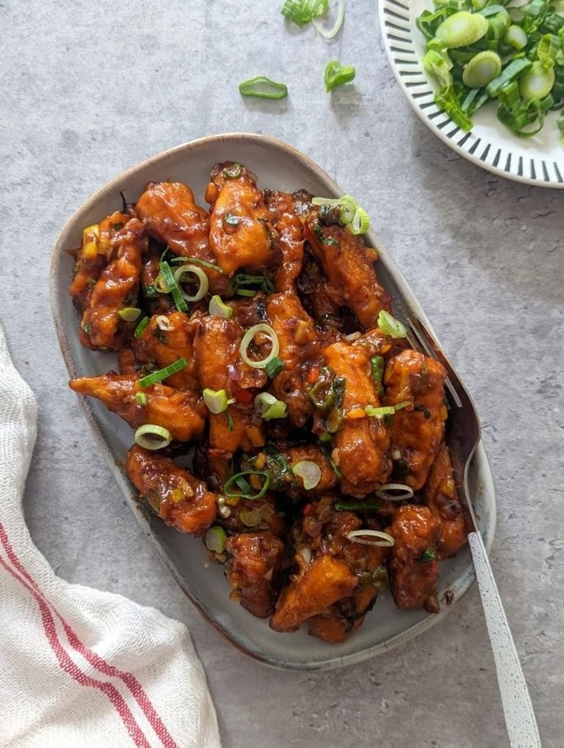 Crispy chili paneer, coated in a rich sauce and garnished with fresh green onions, on a gray plate.